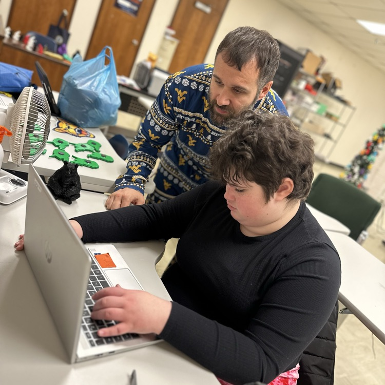 A male teacher wearing blue and yellow-colored pajamas assists a female student with her laptop.