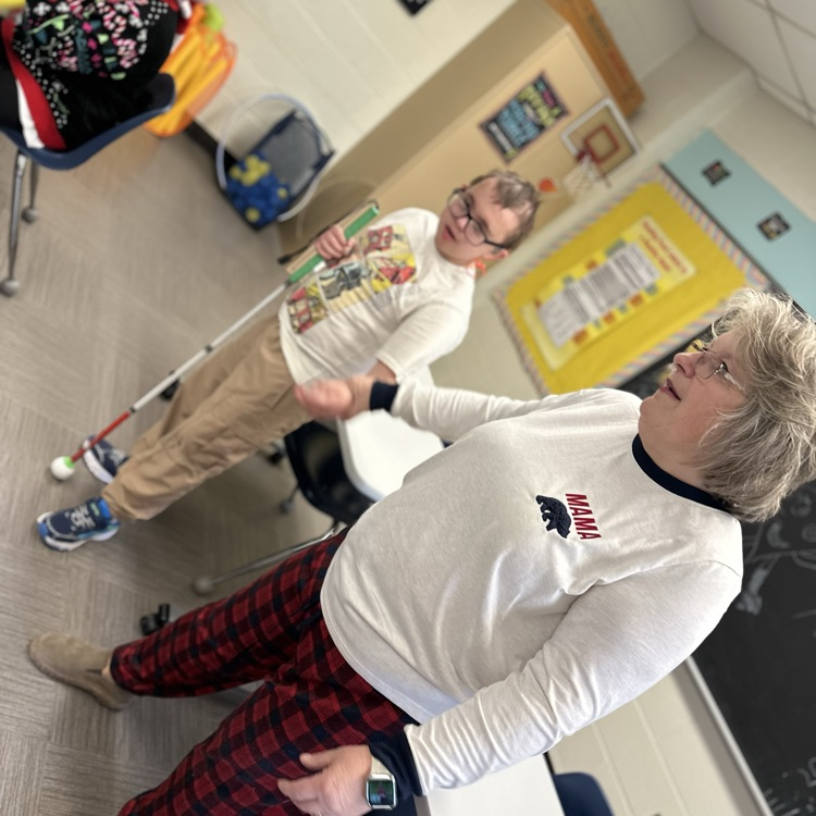 A female aide with short hair and wearing red and black plaid pajama pants holds the hand of a male student. 