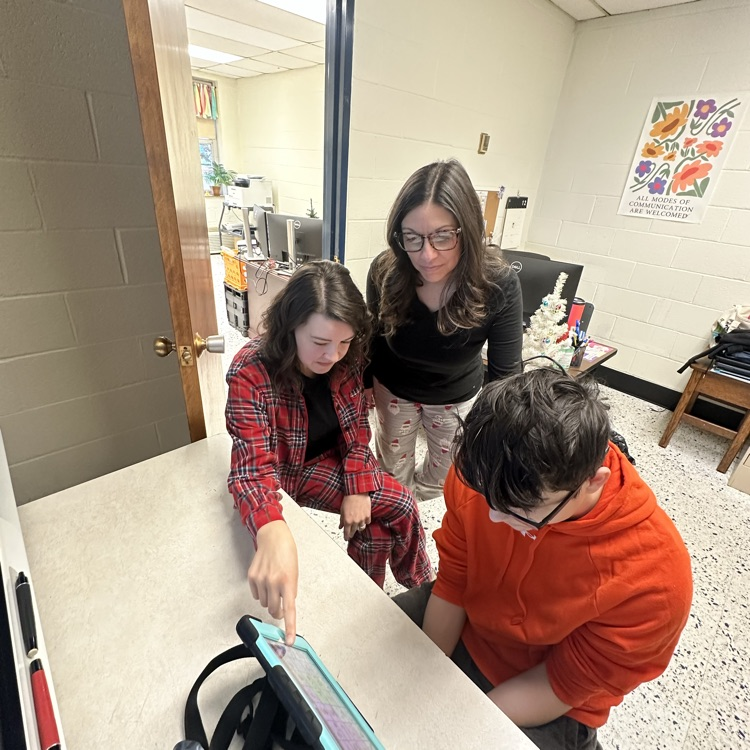 Two female Speech Language Pathologists wearing pajamas assist a male student during his session. 