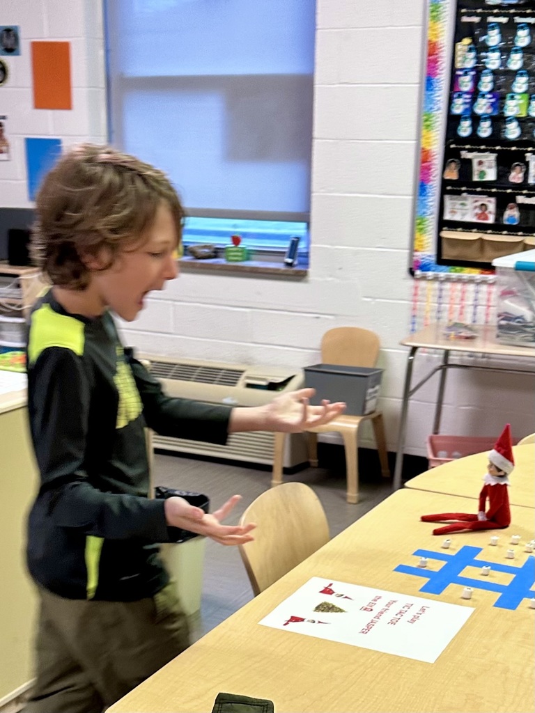 A male student waves his arms with his mouth open as he looks down at the elf that is seated on the table next to the tic tac toe board.