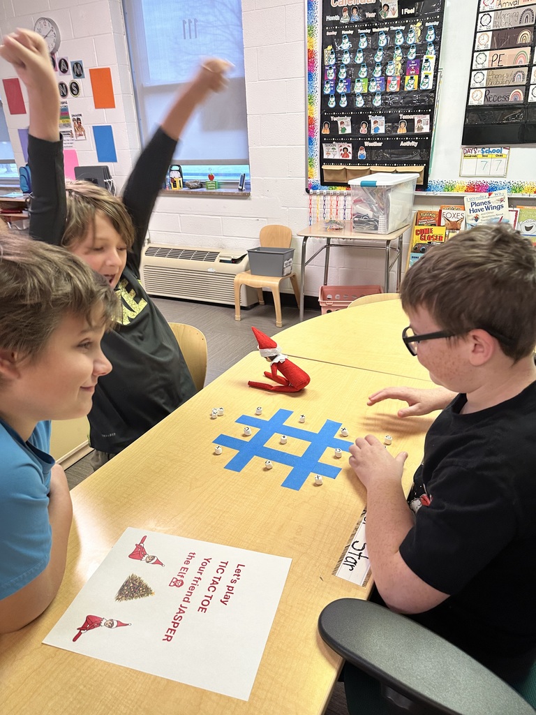 Three male students are seated at a table playing tic tac toe with the elf seated on the table with them. One student has his arms in the air with excitement. 