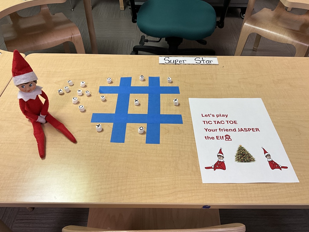 A tic tac toe board made with blue-colored tape. There is an elf dressed in red and white on the left-hand side of the tic tac toe game. On the right-hand side, there is a note from the elf asking to play tic tac toe.