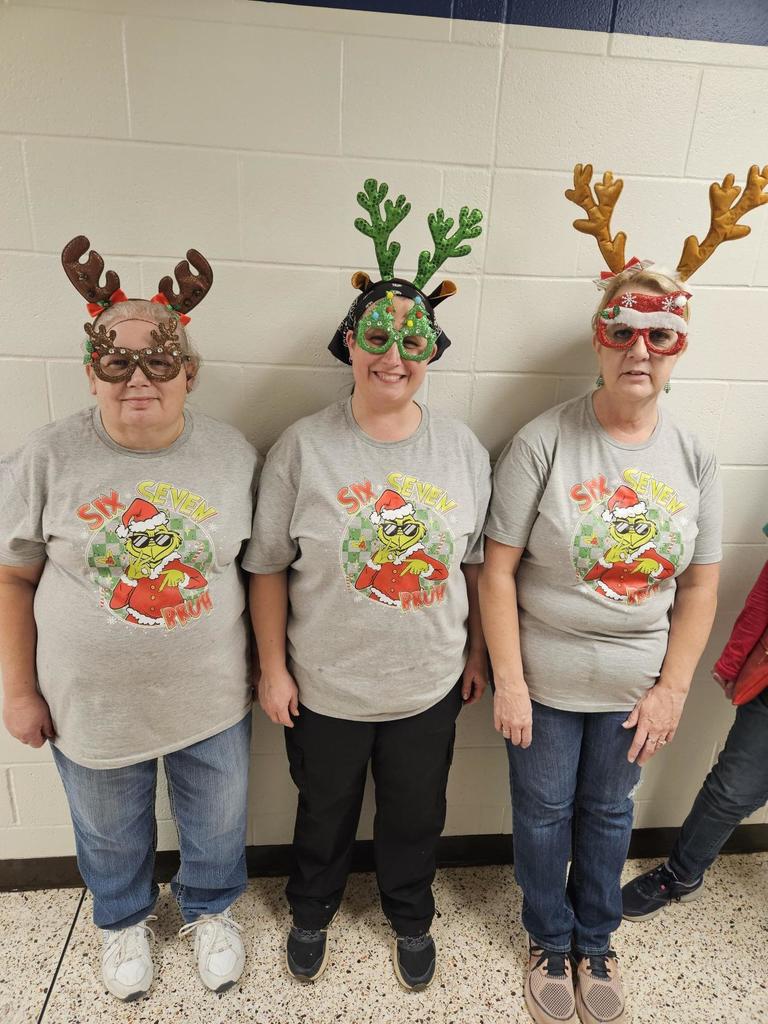 Three female kitchen staff members standing side-by-side wearing matching Grinch shirts and festive headbands.