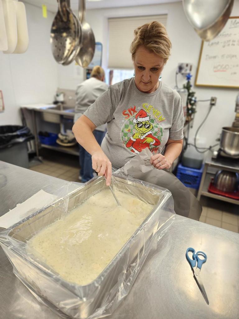 A female kitchen staff member wearing a gray-colored shirt with the Grinch on it, uses a spoon to mix together mashed potatoes that are in a large steel pan.