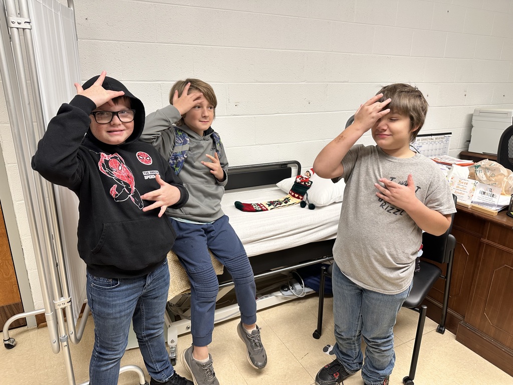 Three male students surround an infirmary bed, where an elf dressed in red and white attire is tucked into a festive sock while laying on the infirmary bed. The three male students use the American Sign Language (ASL) sign for "sick."