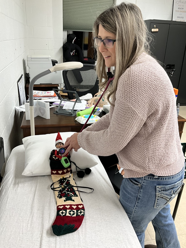 A female nurse, Miss. Tonya, uses her stethoscope on the elf, who is dressed in red and white attire while tucked in a festive sock while lying on the infirmary bed.