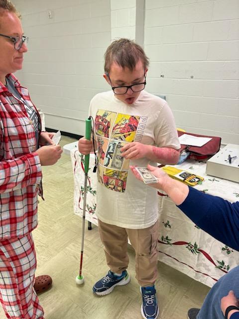 A female aide with red, curly hair and wearing red and white plaid pajamas stands beside a male student wearing a white shirt and glasses. Mrs. Christina Payne is handing them a stack of money.
