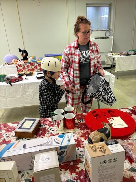 A female aide with red, curly hair wearing red and white plaid pajamas assists a male student wearing a white helmet on his head shop for gifts.