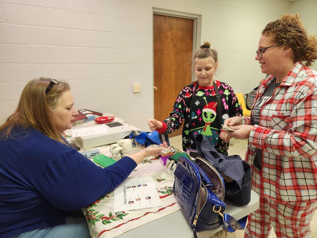 Mrs. Christina Payne, a female wearing a dark blue shirt hands money to a female student wearing a Holiday sweater with a green alien on the front. A female aide with red, curly hair wearing red and white plaid pajamas assists the female student with her money management.