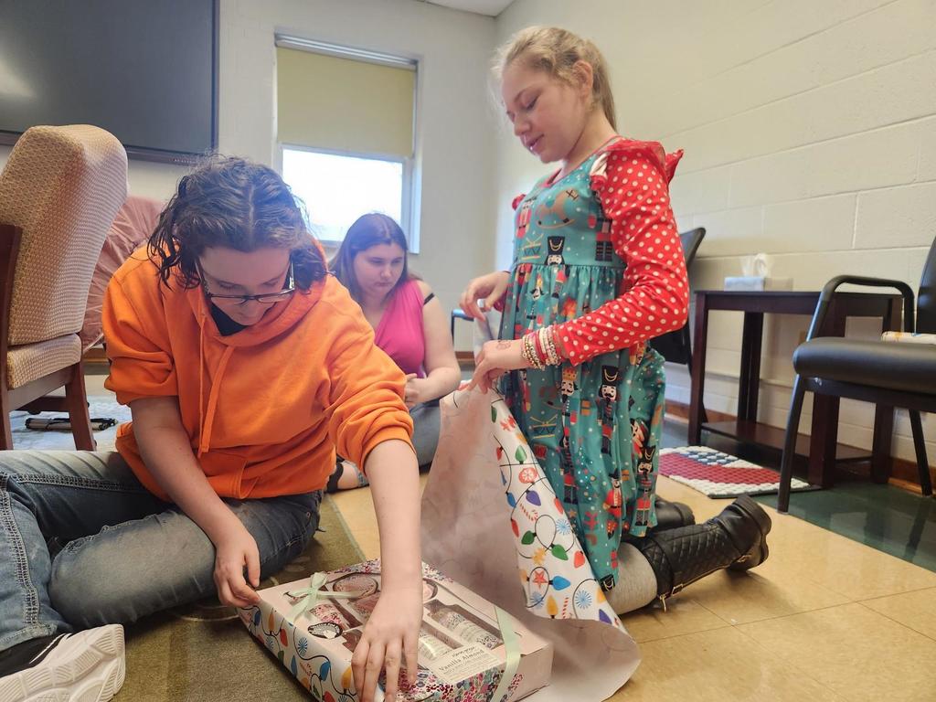 A female CAP student with dark hair and wearing a bright, orange sweatshirt assists a female elementary student with wrapping gifts.