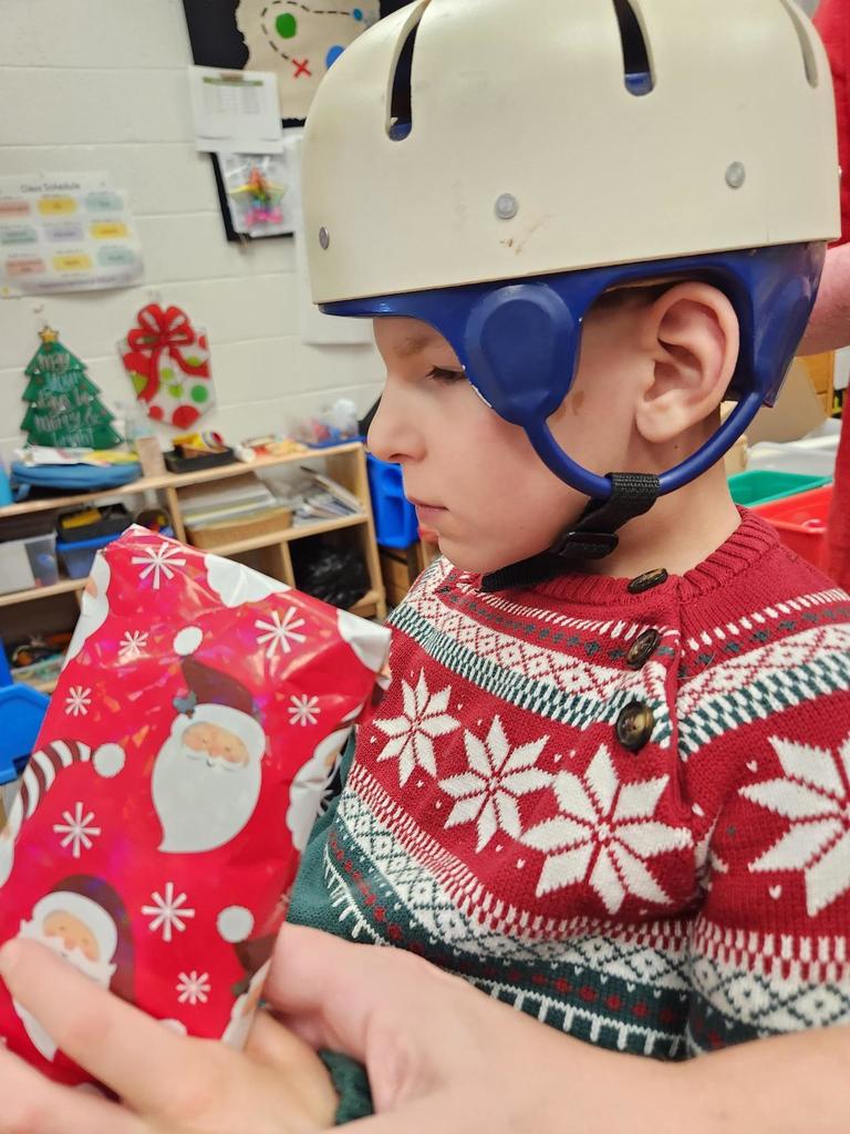 A male student wearing a white helmet on his head and a red and white festive sweater holds up a gift that is wrapped in red and white-colored wrapping paper. 