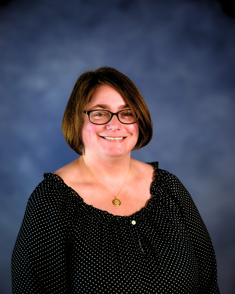 Mrs. Cassie Park. A female with short, brown hair and black glasses smiles for the camera in front of a navy-colored backdrop. She is wearing a black shirt with white dots and a gold-colored necklace around her neck. 