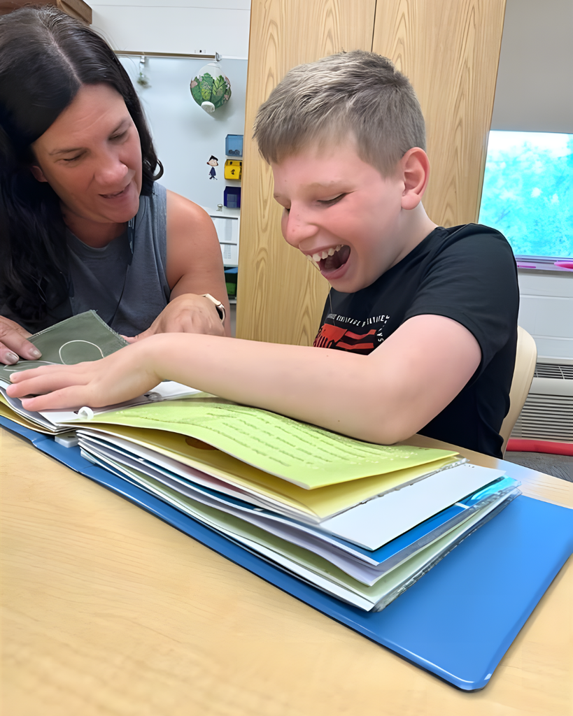 A female with dark, brown hair looks down and smiles as she assists a male student with a book written in Braille. The male student is smiling excitedly as he looks down and touches the Braille dots. 