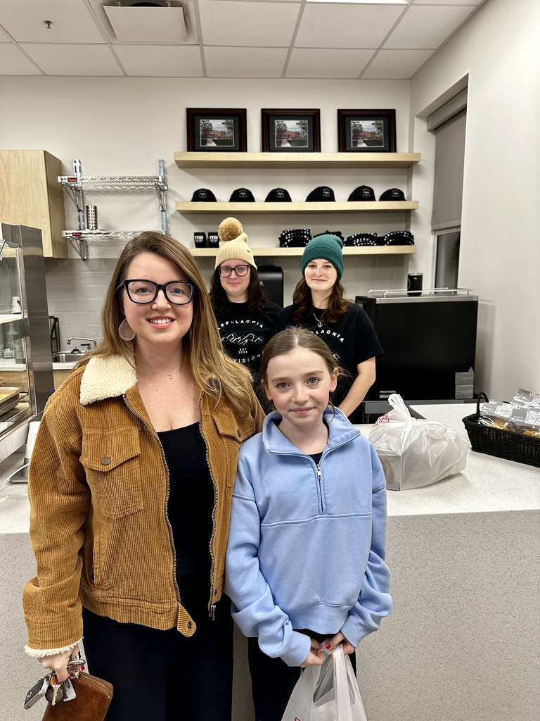 A mother and daughter smile for the camera in the foreground, while in the background behind the cafe counter stands two female-student workers. 