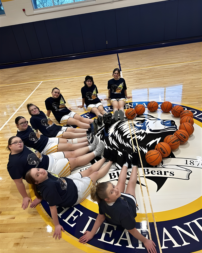 A group of female basketball players dressed in matching navy t-shirts and basketball shorts sit side-by-side on top of the WVSDB Black Bear logo with basketballs sitting on the opposite side of the players. 
