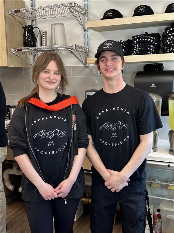 Two students stand side by side while wearing matching Appalachia Provisions t-shirts smile proudly for the camera. The female on the left-hand side has the red-colored ribbon wrapped around her.