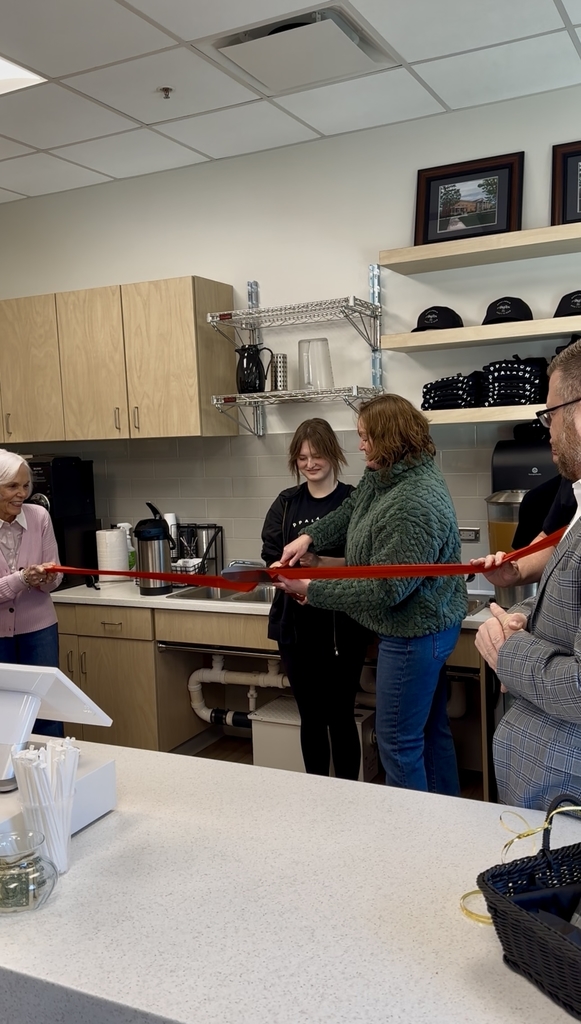 Two individuals stand on either side holding a red-colored ribbon. Two females stand in the center holding a large pair of scissors preparing to cut the red ribbon.