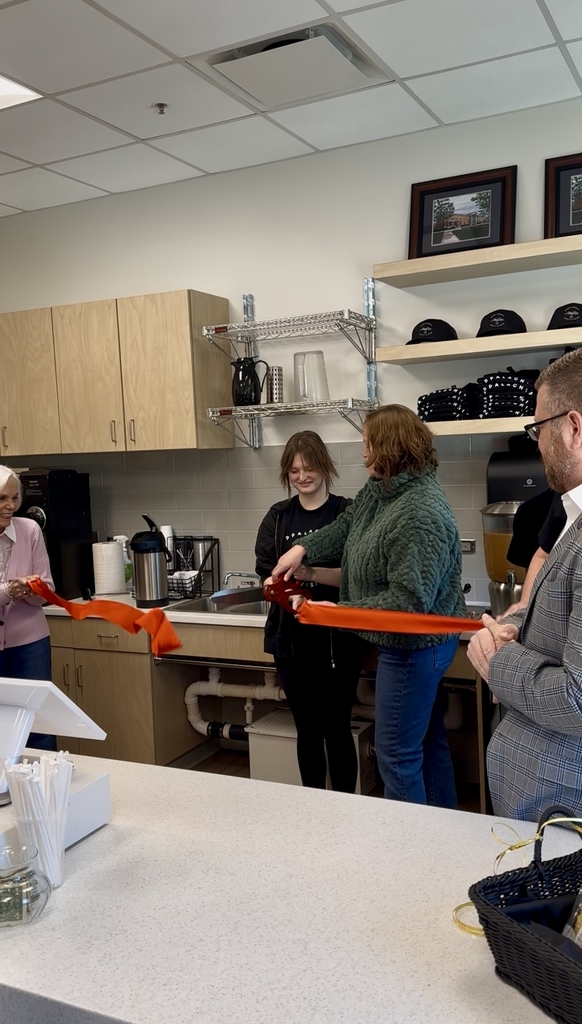 Two individuals stand on either side holding a red-colored ribbon. Two females stand in the center holding a large pair of scissors to cut the red ribbon.