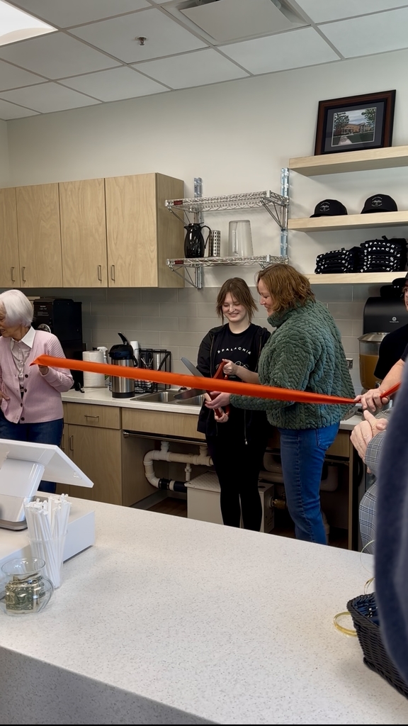 Two individuals stand on either side holding a red-colored ribbon. Two females stand in the center holding a large pair of scissors preparing to cut the red ribbon.