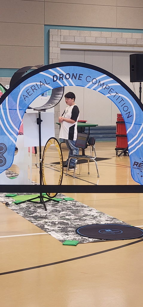 A male student wearing safety glasses and holding up a remote controller used to navigate his drone through the course. 