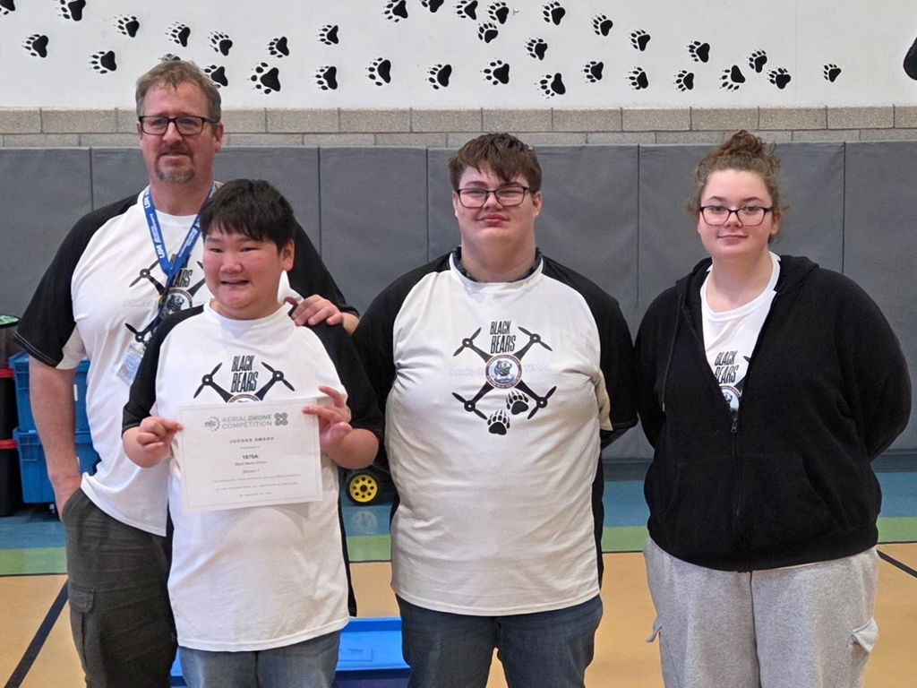 A group of students and their male coach stand side by side for a photo. A male student in the front proudly holds up a certificate of recognition. 