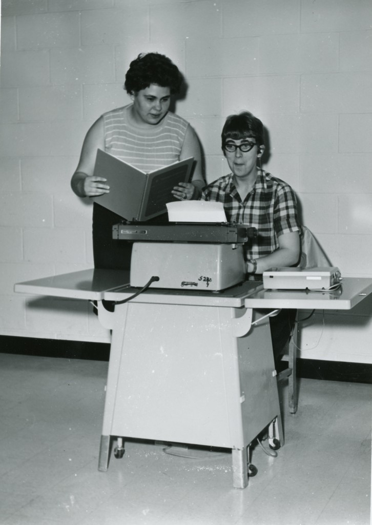 An individual stands to the left while holding a large book, while a seated individual to the right types on a Braille writer.
