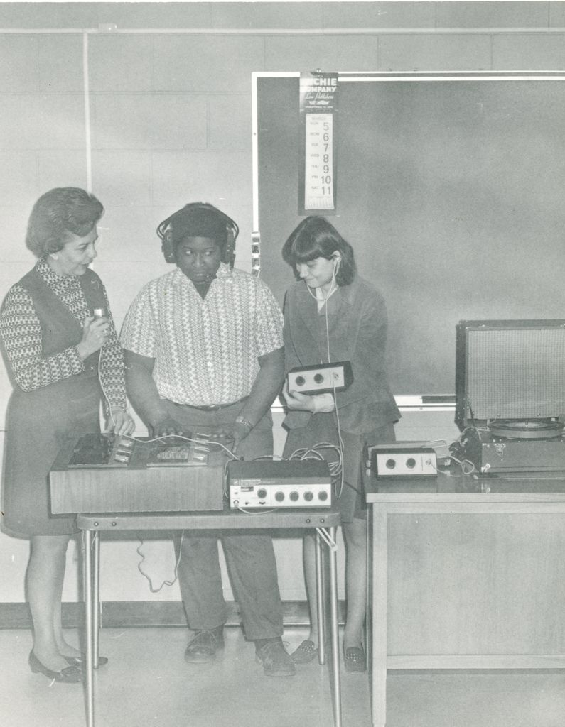 Three individuals standing side-by-side, gathered around a desk with Braille equipment sitting on top. The individual in the middle is wearing headphones and typing on a Braille writer.