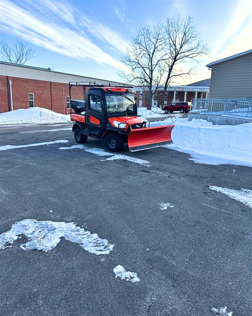 An orange Kubota tractor with a plow on the front is parked in a parking lot with several buildings in the background. There is white snow that has been plowed. 