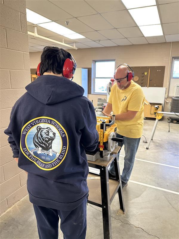A male student wearing a navy-colored sweatshirt with the WVSDB Black Bear logo on the back works with a male instructor wearing a yellow-colored shirt with a machine that smooths wood. 
