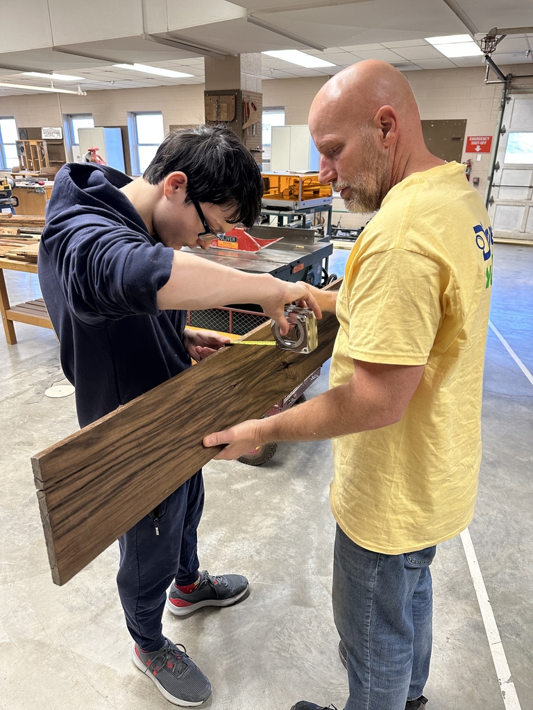A male instructor holds a large piece of brown wood as a male student stands across from him using a measuring tape to measure the width of the piece of wood. 