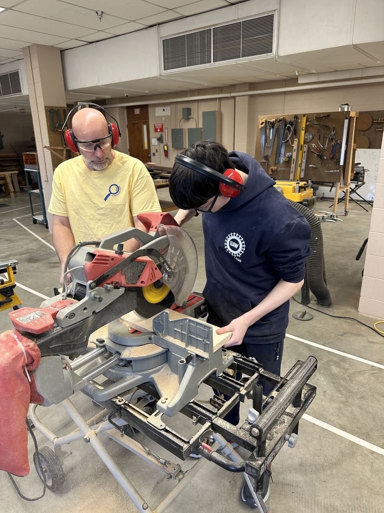 A male instructor wearing a yellow-colored shirt, hearing and eye-protective equipment assists a male student who is also wearing hearing and eye-protective equipment use a table saw to cut a piece of wood. 