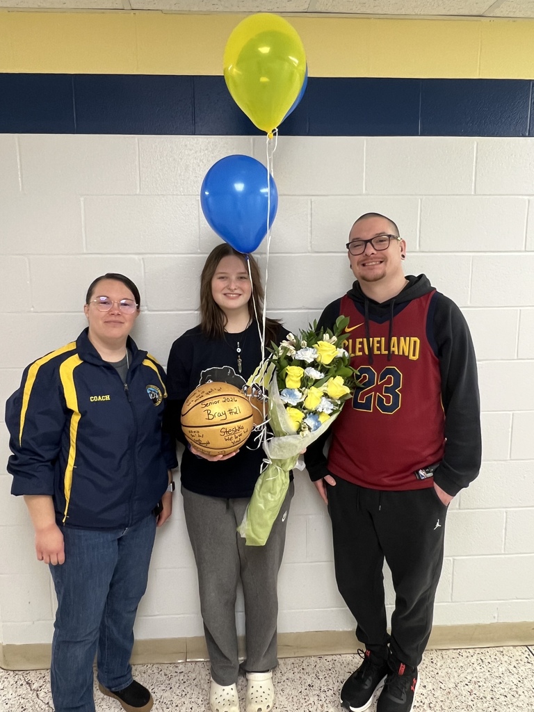Two coaches stand on either side of a female basketball player who is holding a basketball with team members signatures, a bouquet of flowers, and blue and yellow balloons. They are all smiling proudly at the camera. 