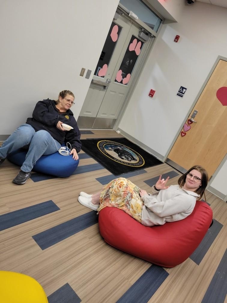 A female Residential Care Specialist and a female residential student are seated on bean-bag chairs. The female student holds up the "I love you" sign in American Sign Language (ASL).