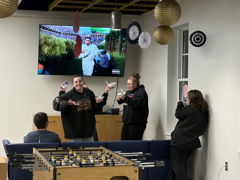 Students mingling with one another in the student union. The Super Bowl is playing on a TV behind the students. 