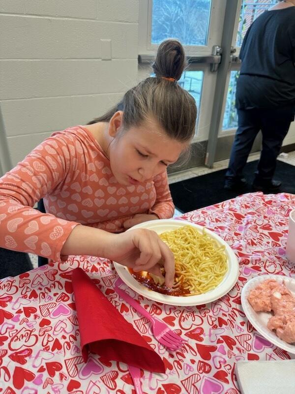 A female student seated at a table with tablecloth with red and pink hearts enjoys her pasta meal.