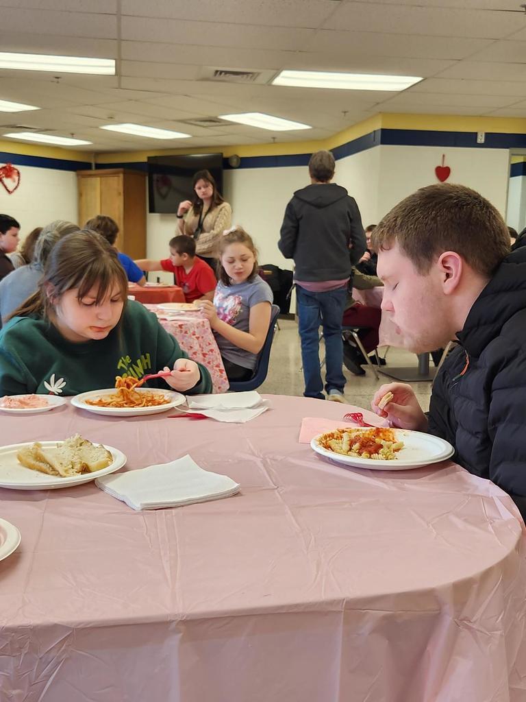 Students seated at a circular table with a pale pink tablecloth on top, while enjoying their pasta meal.