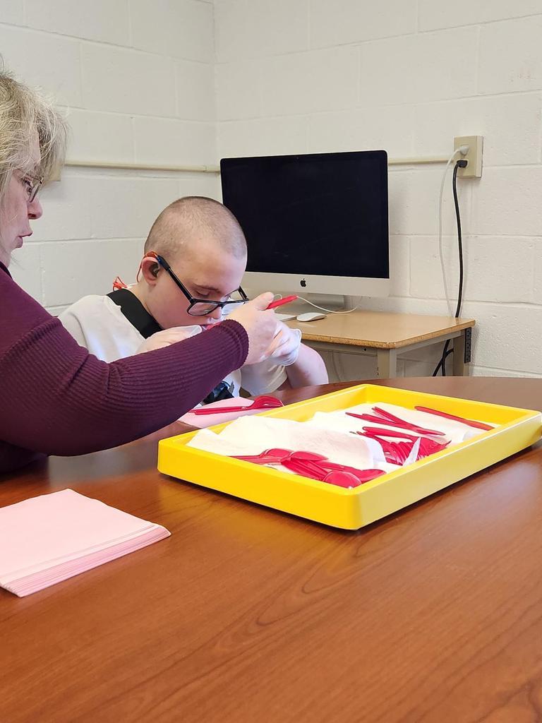 A male student looks down and puts together utensils and napkins with the help from a female aide sitting beside him.