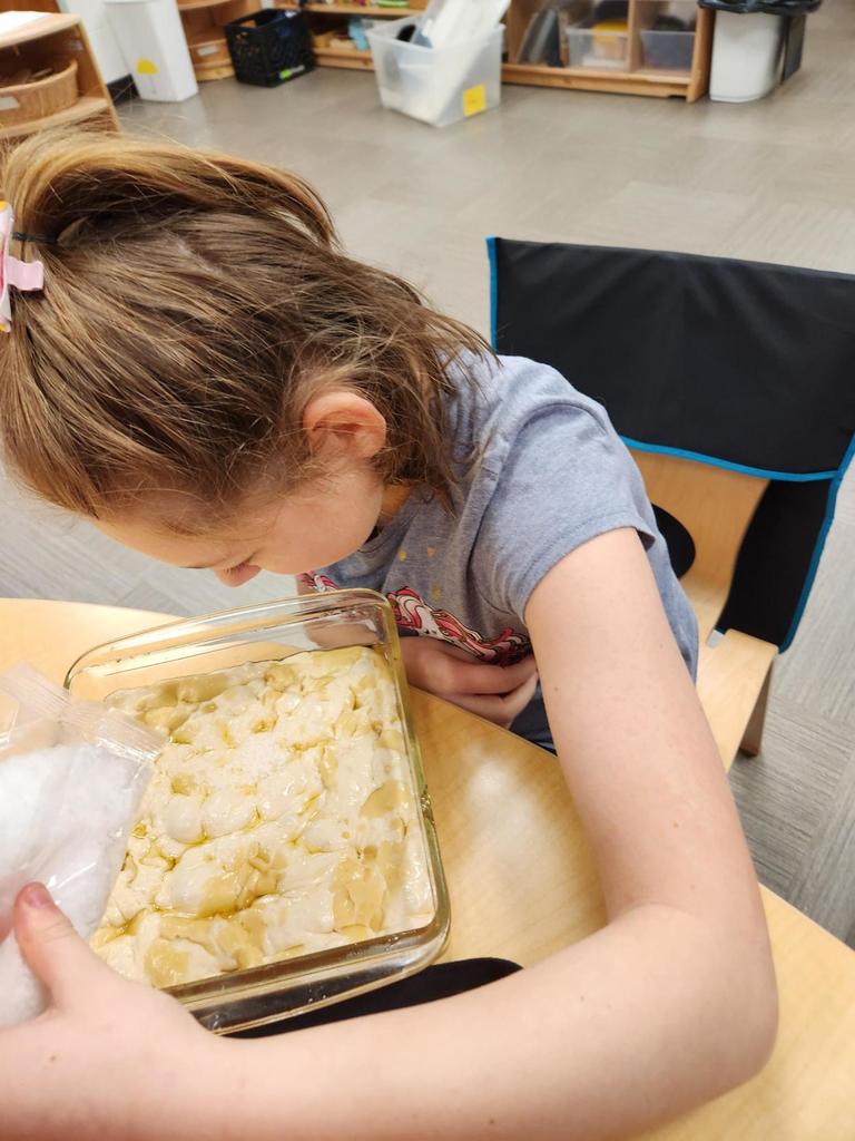 A female student seated at a table has assistance with prepping focaccia bread. 