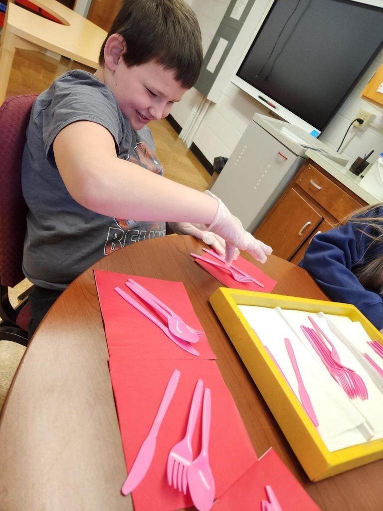 A male student is seated at a table as he looks down to put together utensils and napkins.