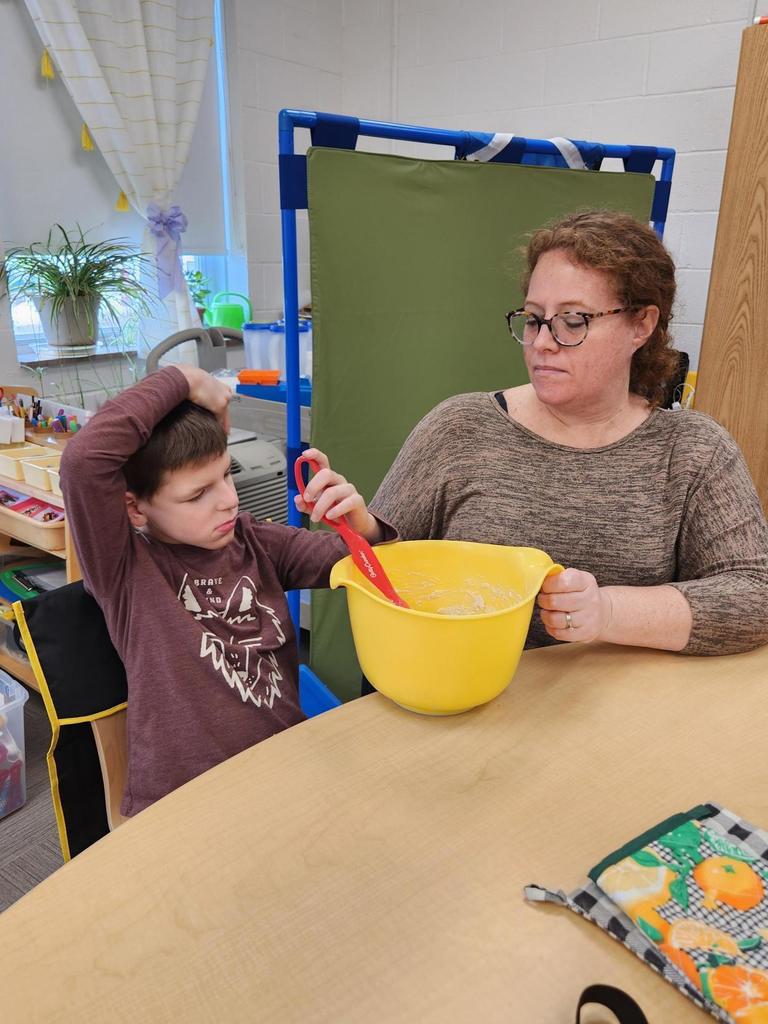 A male student uses a red spoon to stir contents that are in a large, yellow bowl. He has assistance from a female aide sitting beside him.