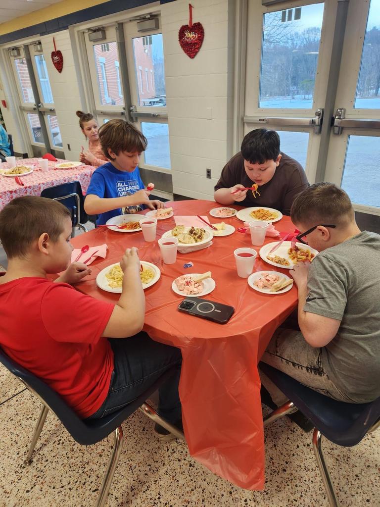 Male students seated at a circular table with a red tablecloth on top look down as they enjoy their pasta meal.