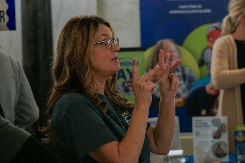 A female with long hair and wearing glasses signs American Sign Language (ASL) with her hands. 