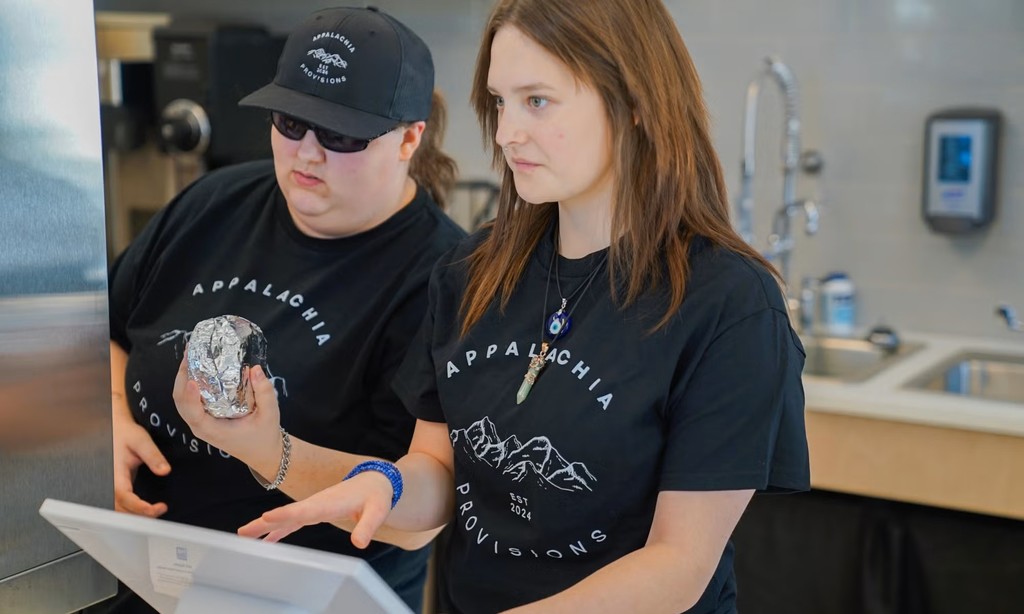 Two female students wearing matching black shirts with the Appalachia Provisions logo on the front. They are working the cash register in the cafe.