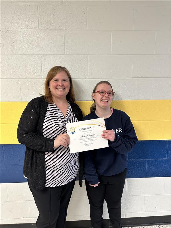 Mrs. Hesse, Principal of WVSDB stands alongside, Mira, a female student. They are both holding up a certificate together. They are standing in front of a white wall with blue and gold stripes in the center of the wall. 