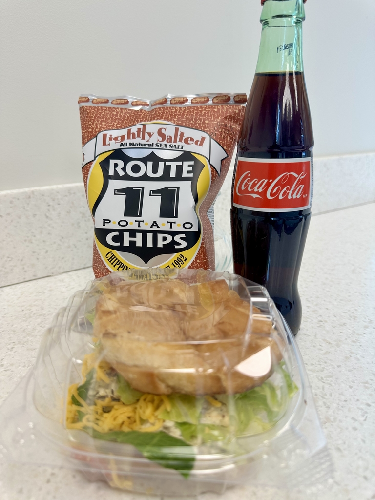 A Chicken Bacon Cheddar Croissant with a bag of chips and a Coca-Cola in a glass bottle in the background behind the croissant. 