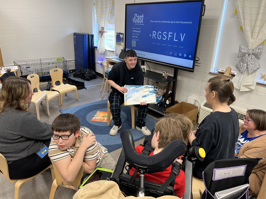 A male high school student seated in a chair in front of the elementary class holds the book open for the students to see the illustration.