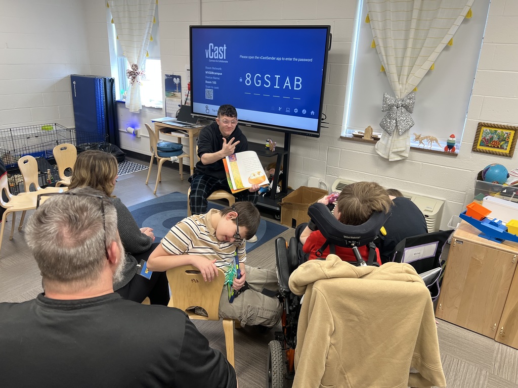 A male high school student is seated in a chair in front of elementary students gathered around him. The high school student holds a book in one hand and signs in American Sign Language (ASL) with the other hand. 