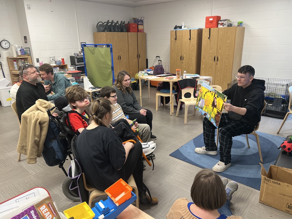A male high school student seated in front of an elementary class holds a book open wide so the students can see the illustration. 