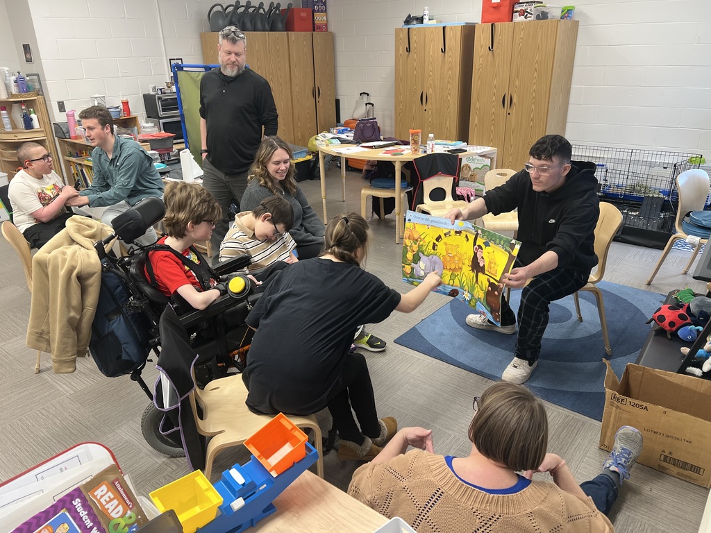 A male high school student seated in front of an elementary class holds a book open wide so the students can see the illustration. 