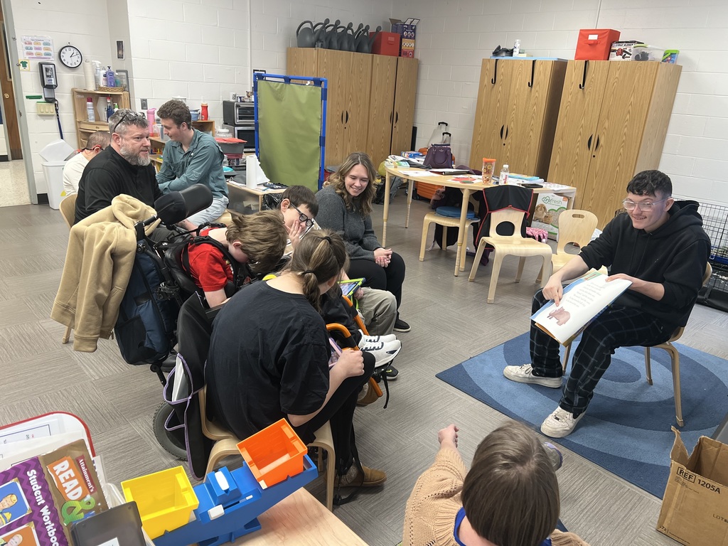 A male high school student is seated in front of an elementary class holds a book with one hand and points to an illustration with the other hand. 